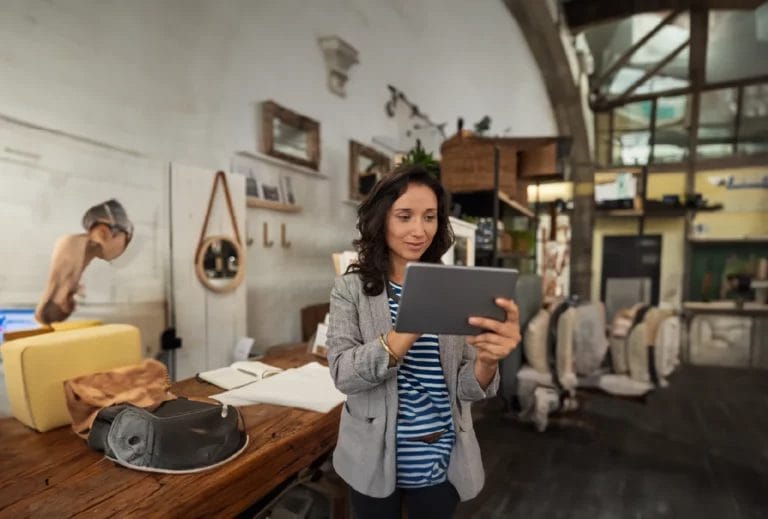 A woman wearing a navy-and-white stripe top and gray blazer holds a business tablet and smiles in a store decorated in cream and brown tones.