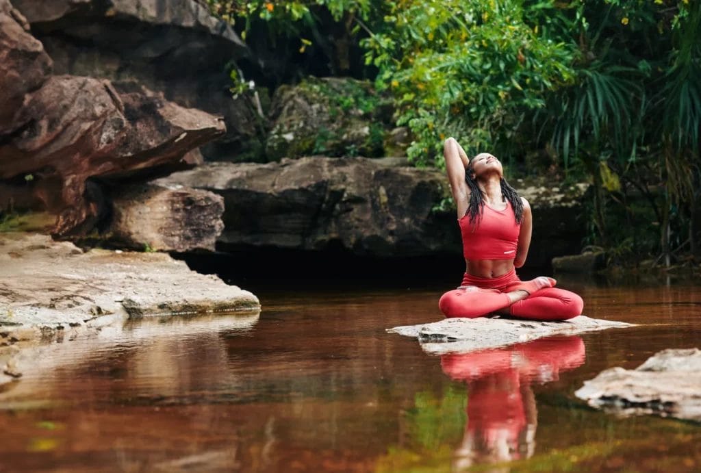 A yogi with black dreadlocks and red athletic clothes performs a variation of lotus pose while sitting on a rock surrounded by water, other rocks, and green plants.