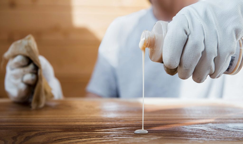 A carpenter is pouring wood polish onto an antique wooden surface as part of the restoration process. If the polish was not made correctly, it could damage the piece and product liability insurance may help pay for the damages.