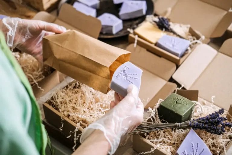 A soap maker wearing a green apron and clear plastic gloves packs a bar of lavender soap into a brown paper bag above multiple cardboard boxes prepped for shipping.