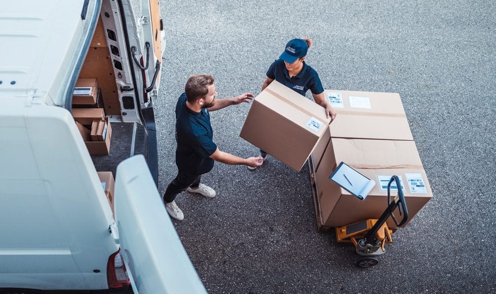 An employee is passing boxes off a dolly to another employee who is loading them into the back of a van.