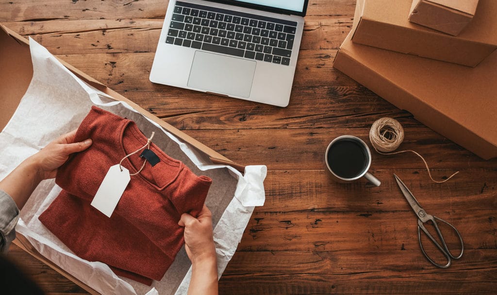 A small business owner is working on a wood table as they package up a red shirt into a box to be delivered to a customer.