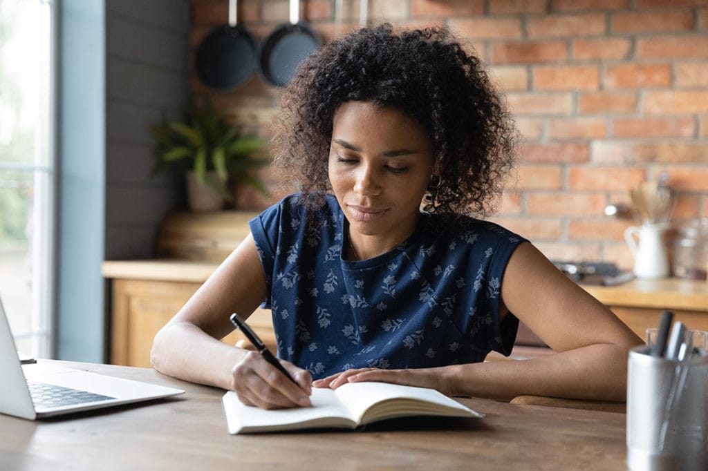 A woman sitting at her kitchen table in front of a laptop taking notes in a journal.