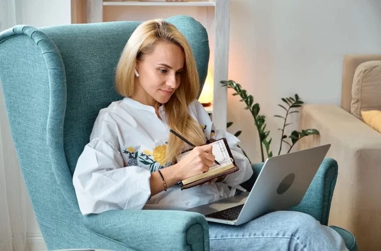 Woman sitting on a chair using a laptop and writing in a notebook.