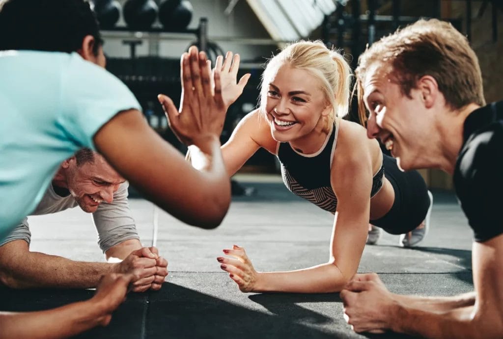 Two women high-five while planking with others in a circle at the gym