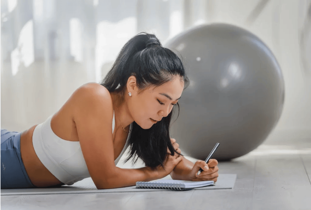 A young woman writing in her notebook while on her yoga mat with an exercise ball in the background.