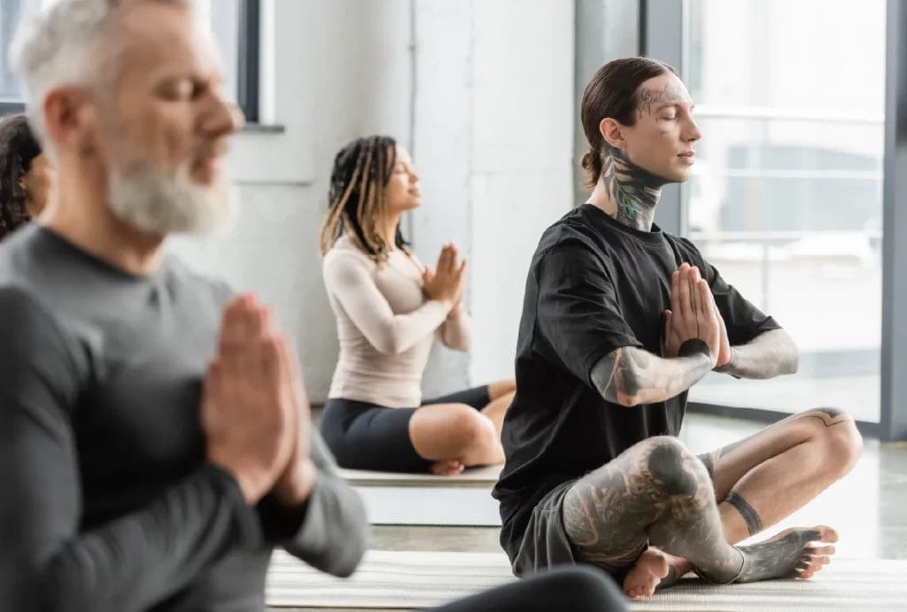 A tattooed man practices anjali mudra in yoga class with other students.