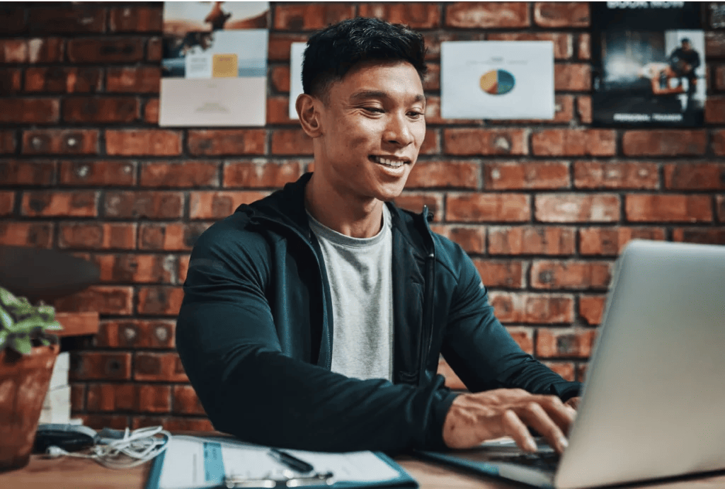 A male fitness entrepreneur wearing a gray T-shirt and teal hoodie smiles while working on a laptop at the gym reception desk.