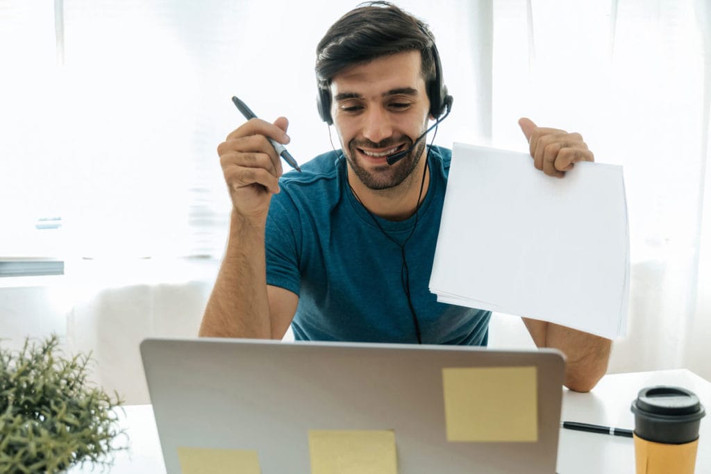 A tutor giving a lesson, wearing a headset sitting at his desk in front of a laptop holding up paper and a marker