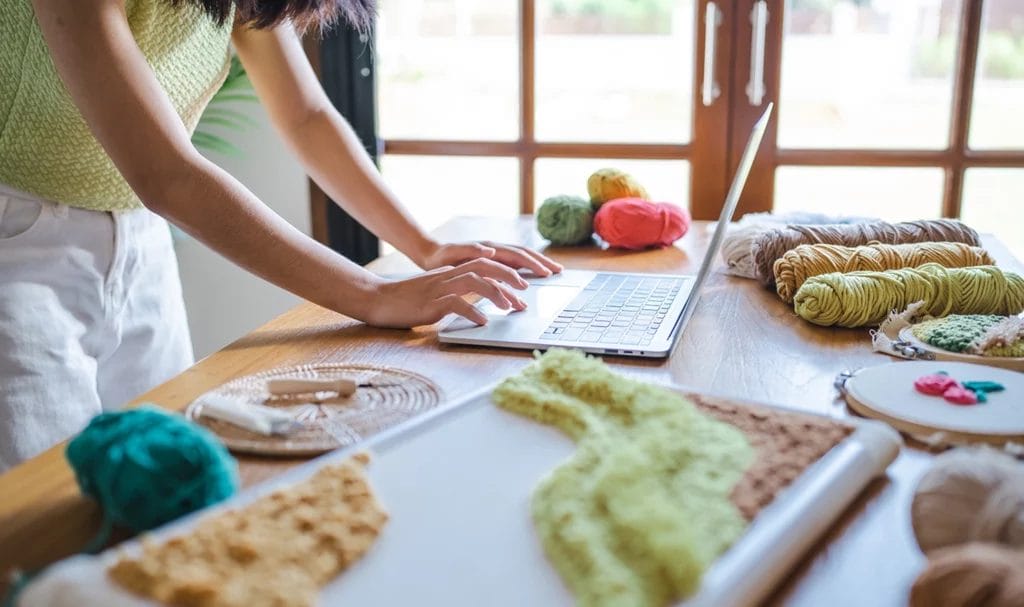 A business owner is working on a craft table in a home art studio on a laptop, applying for a product insurance quote online.