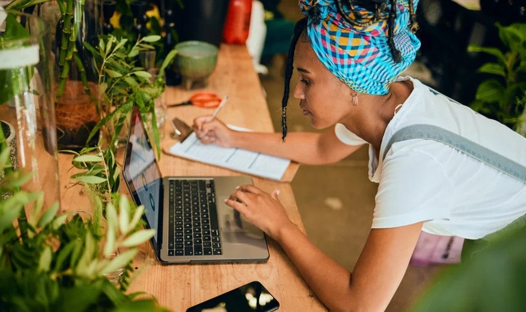 A young woman is working in her small business floral shop as she leans down to work on a laptop on a desk. Getting small business product liability insurance will aid her in her business planning process.