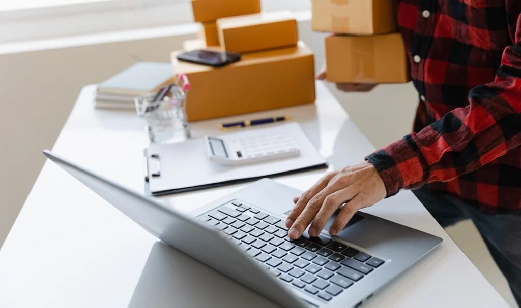 A business owner's hands are working on a laptop on a table next to products ready in packages to be shipped. Product insurance for small business can help to protect them from claims that arise from these products.