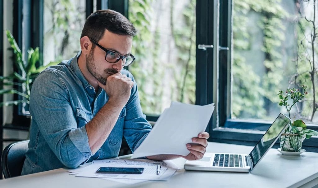 A young business owner is looking over paperwork in his home office next to his laptop as he decides on what product liability insurance to buy.