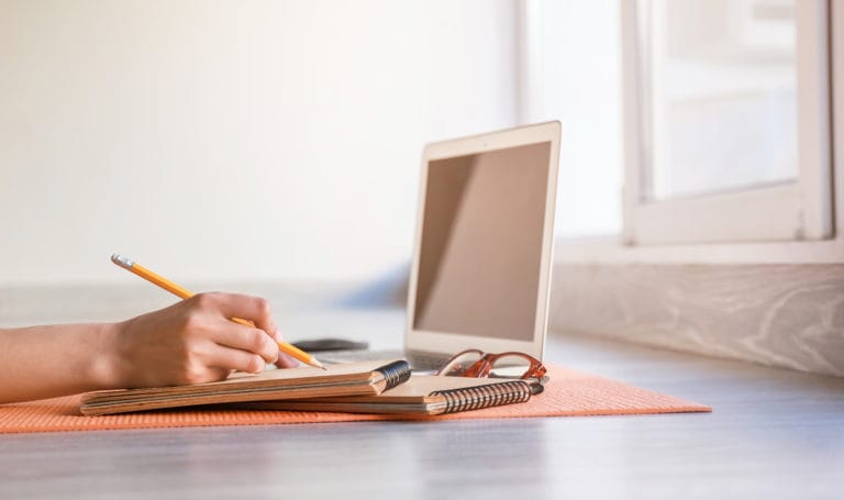 A side view of a person's hands writing on a notebook next to their laptop on a yoga mat as they work on a personal training business plan.