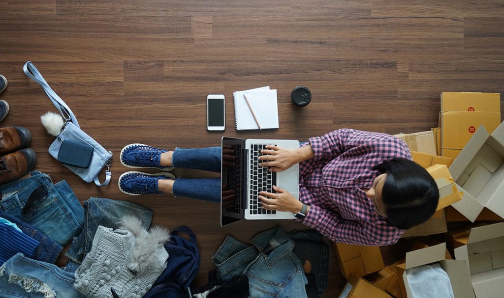 A business owner sits on the ground as they work on a laptop next to boxes and products their business sells.