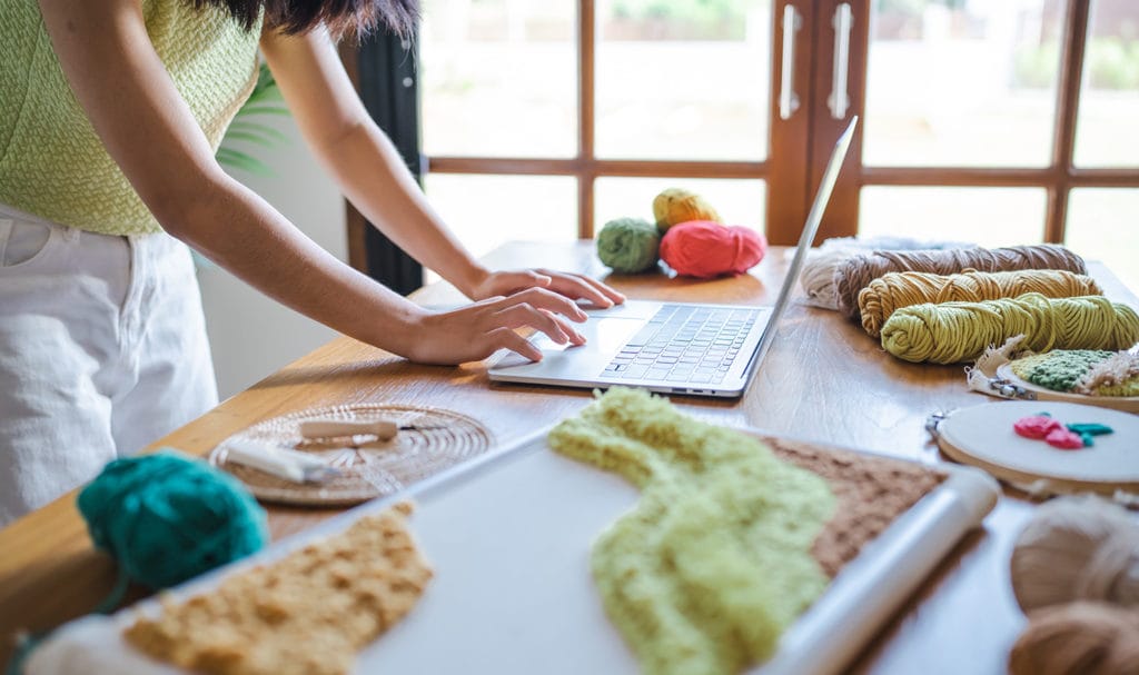 A business owner is working on a craft table in a home art studio on a laptop, applying for a product insurance quote online.