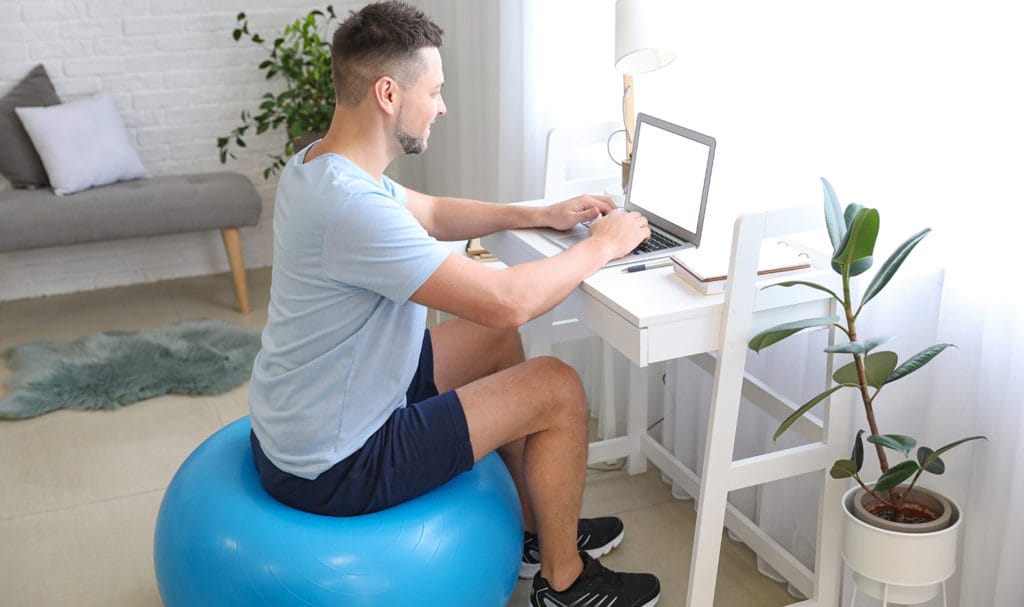 A personal trainer sits on a blue yoga ball inside his home office as he works at a desk on his laptop to create facebook ads for personal trainers so he can better promote his business online.