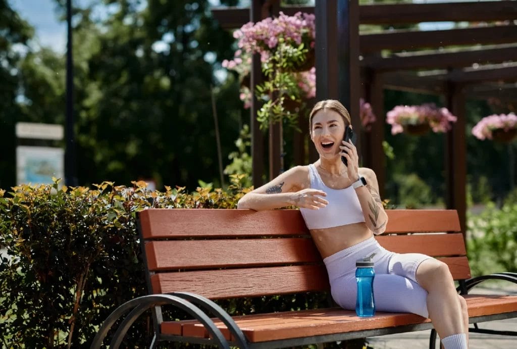 A personal trainer with vitiligo talks on the phone while on a park bench.