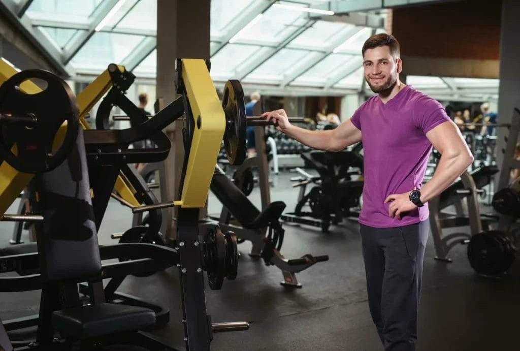 A personal trainer wearing a purple tee and black pants smiles while standing next to a large weightlifting machine.