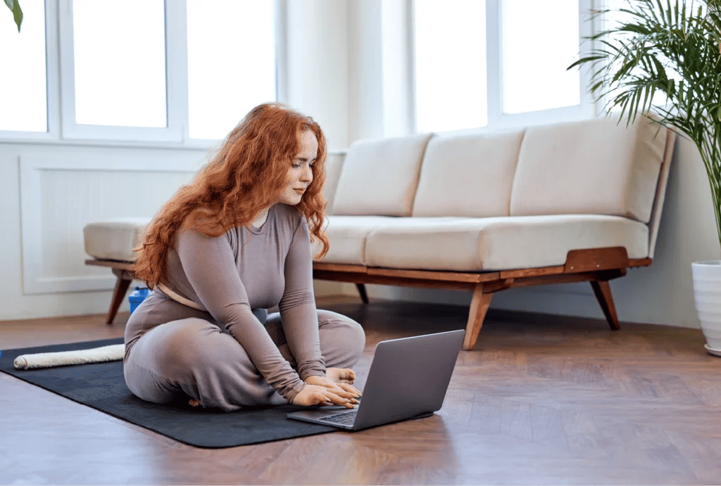 A personal trainer with long curly red hair sits cross-legged on a mat while typing on the computer.