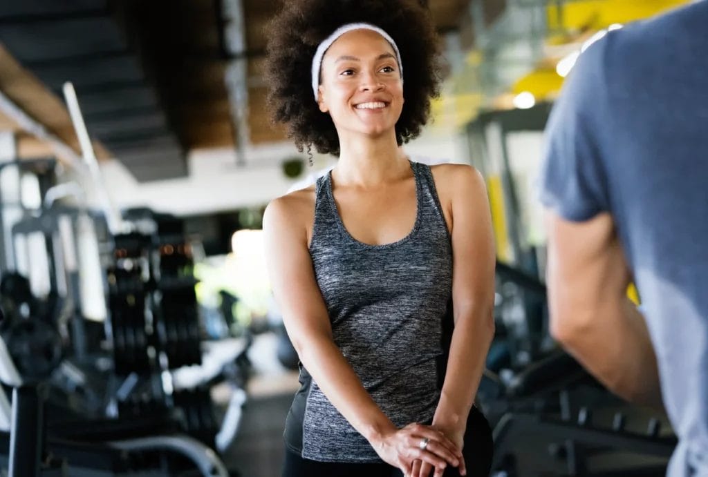 Woman with an afro, wearing a dark gray tank, smiles in front of a blurred gym background.