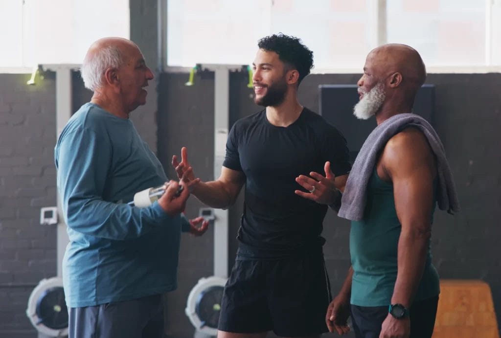 A personal trainer with a beard and curly hair, wearing all black, talks to two older men in front of a set of erg machines.