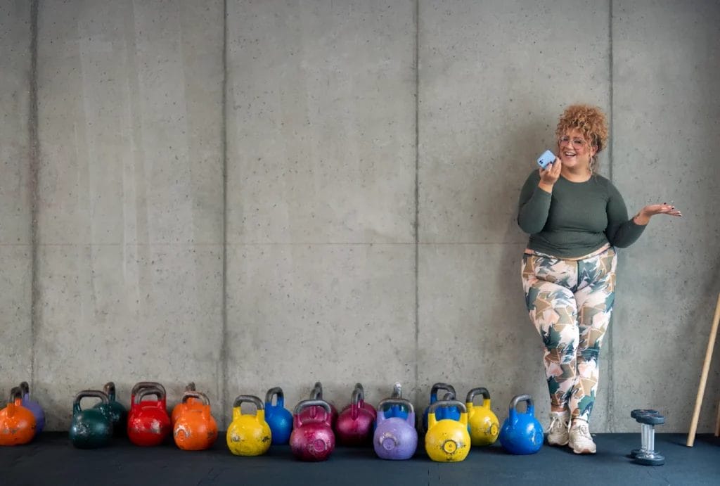A fitness client with curly hair and glasses talks on the phone while standing next to a collection of colorful kettlebells.