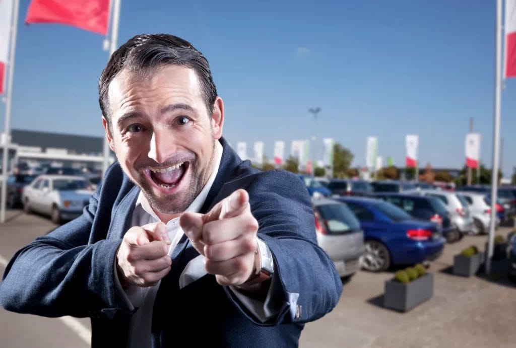 An exuberant man wearing a suit in front of a car lot points at the camera with both hands while smiling wide.