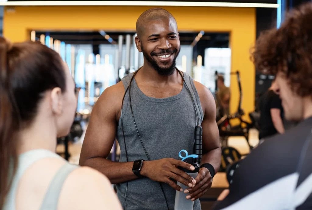 A trainer with a beard and mustache, wearing a gray tank, talks to two potential clients.