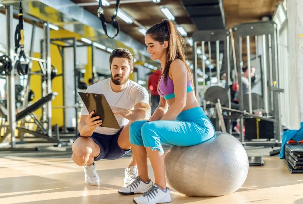 A trainer with a beard crouches next to a new client, pointing out a plan on a clipboard while the client sits on a yoga ball, smiling.