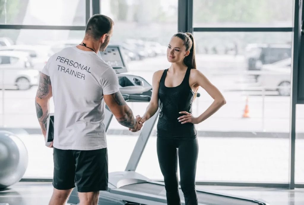 A tattooed personal trainer wearing a personal trainer tee greets a new client wearing black athletic clothes.