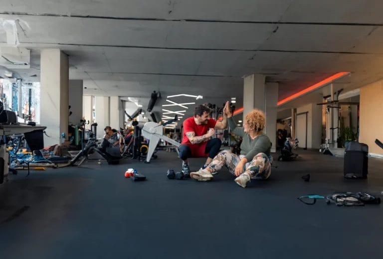 A tattooed trainer in red athletic wear high-fives a new client as she sits on the ground.