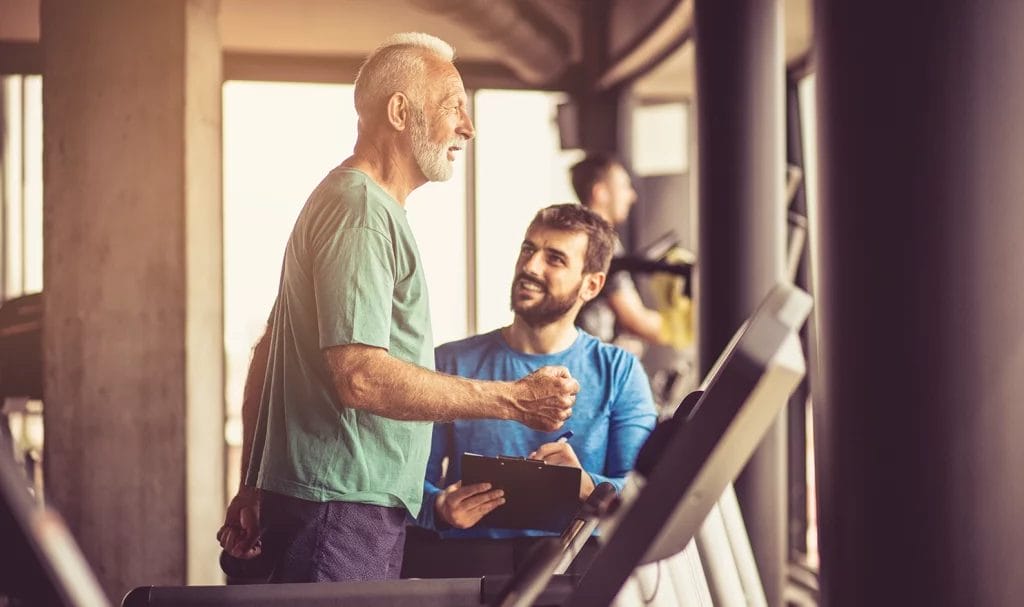 Male fitness trainer with a clipboard talking to an older male client walking on a treadmill.