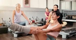A Pilates teacher demonstrates a pose on a reformer machine while students observe from their own machines during a class.