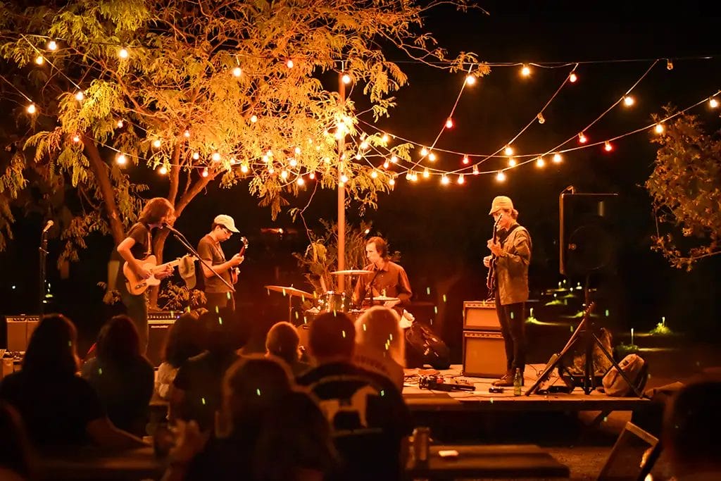 A band of four musicians plays on a small stage at an intimate outdoor venue at night with string lights forming a canopy over the stage.