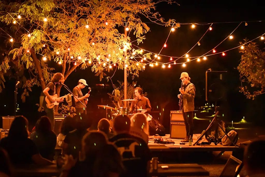 A band of four musicians plays on a small stage at an intimate outdoor venue at night with string lights forming a canopy over the stage.
