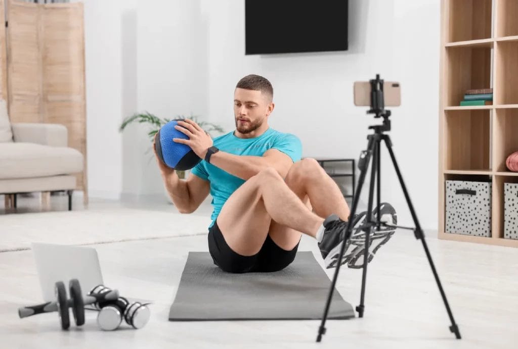 A personal trainer is on a yoga mat doing core workouts with a medicine ball in front of a smart phone on a tripod and a laptop as he livestreams his session for clients