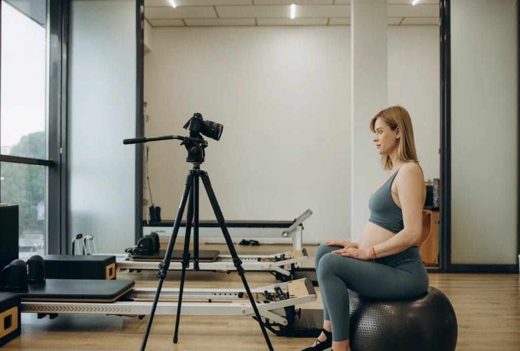 A personal trainer is sitting on an athletic ball in front of a camera, ready to film a training session