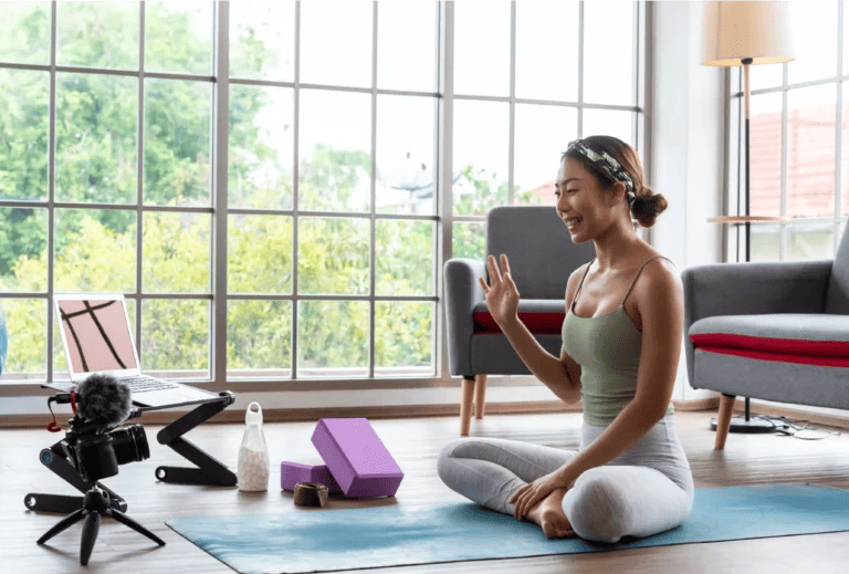 A virtual personal trainer sitting on a yoga mat in their home waving hello to a camera and laptop in front of them