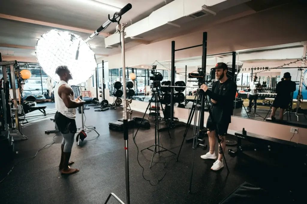 Man recording a personal trainer giving exercise instructions in a gym with lights and recording equipment.