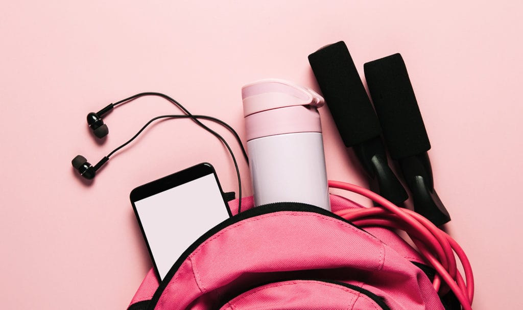 A pink backpack filled with workout gear like headphones, a water bottle, and a jump rope, are displayed on a pink background.