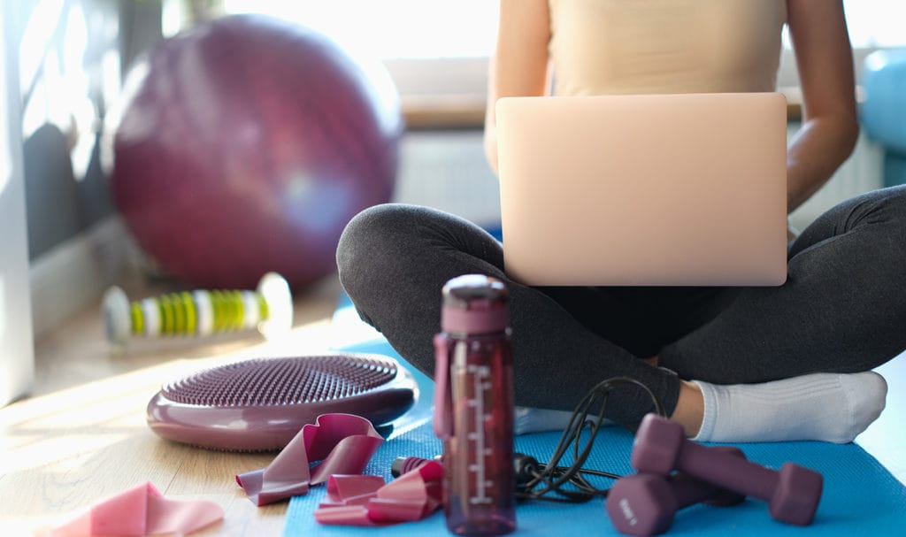 A fitness instructor sits on the floor next to their gear while they work on their laptop.