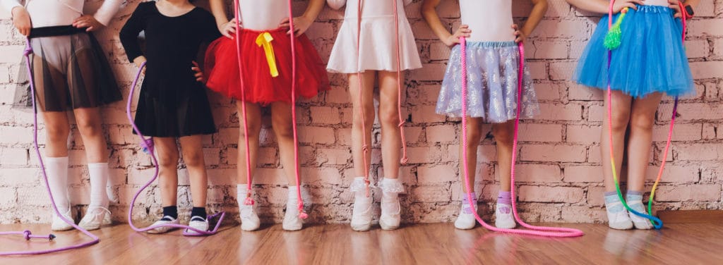 Kids in ballet outfits stand in a row by a wall with jump ropes.