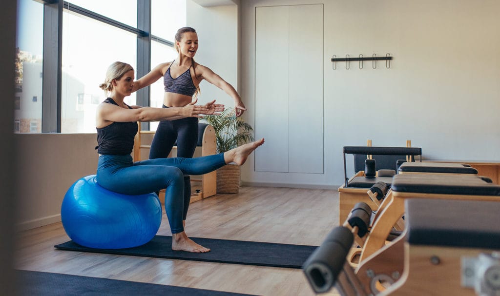 A pilates teacher helps a client strengthen their core on an exercise ball.