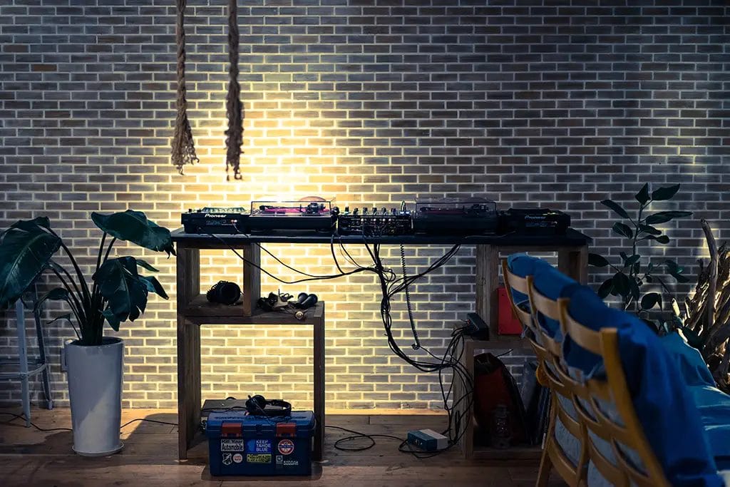 A table covered in DJ equipment including turntables and microphones surrounded by large potted plants in front of a brick wall.