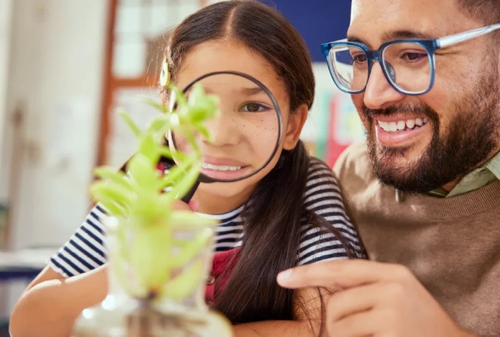 Happy tutor or teacher and elementary school student looking at plant under a magnifying glass