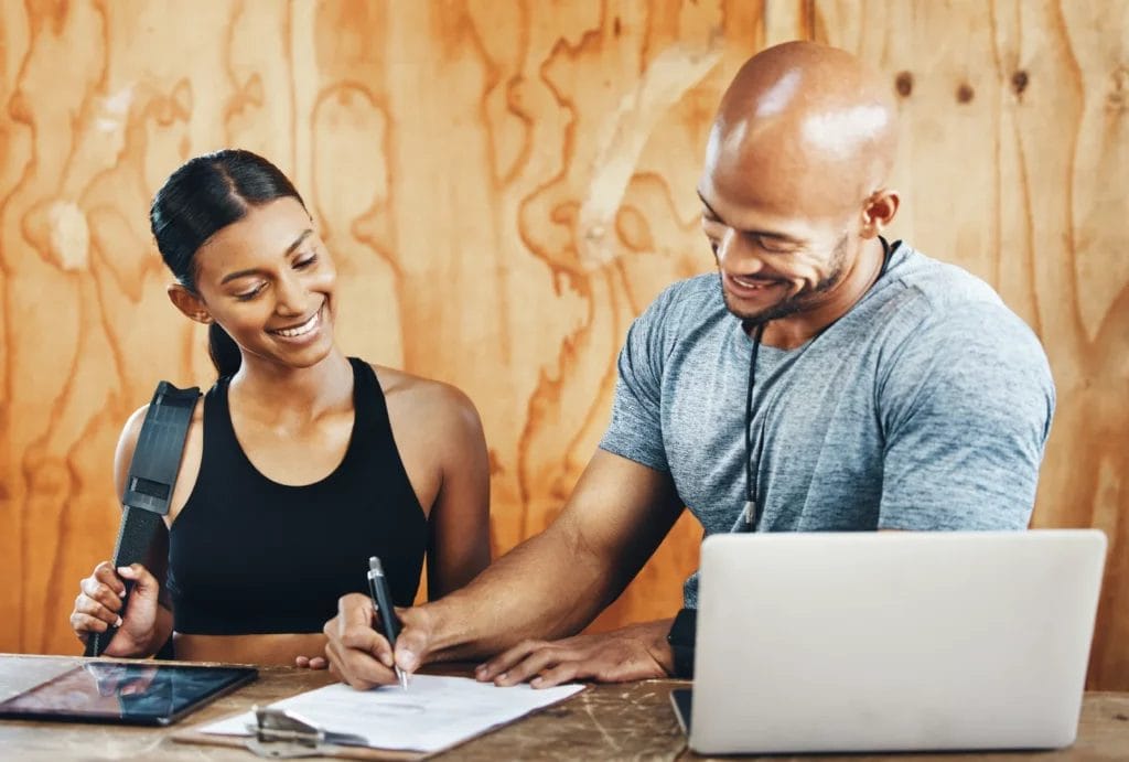 A personal trainer wearing a gray T-shirt signs a form with a client wearing a black top and a backpack in front of a wooden desk with a laptop and a tablet on it.