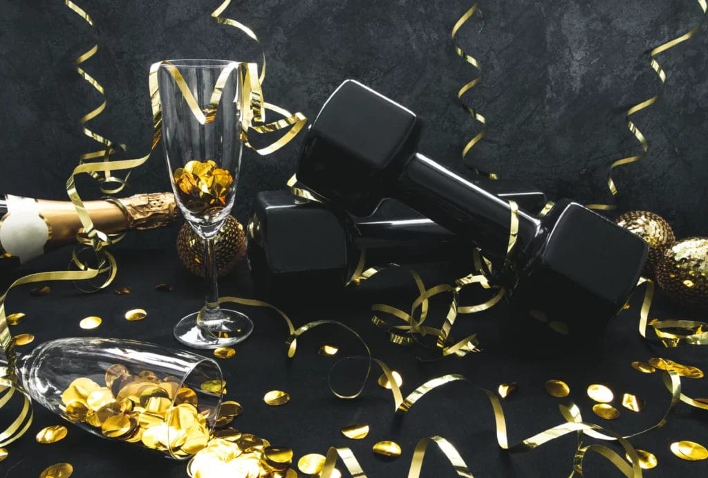 A pair of black dumbbells with champagne flutes, a champagne bottle, and gold confetti and ribbon on a dark gray backdrop.