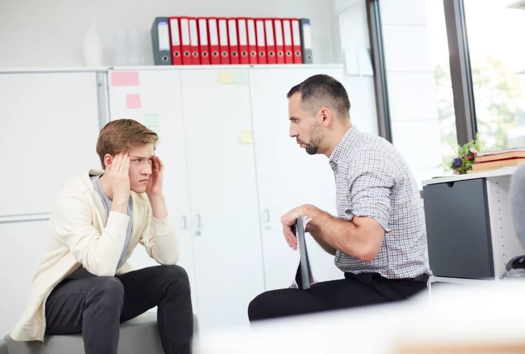 A tense teenager listens to advice from a coach inside a classroom.