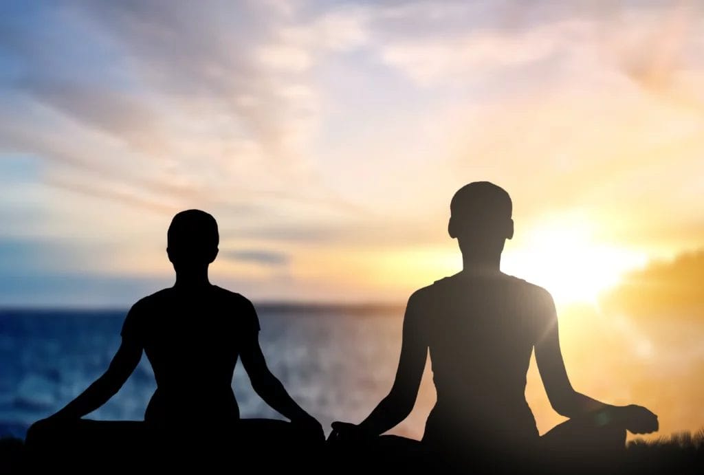 Two people silhouetted against a sunrise meditate by the ocean.
